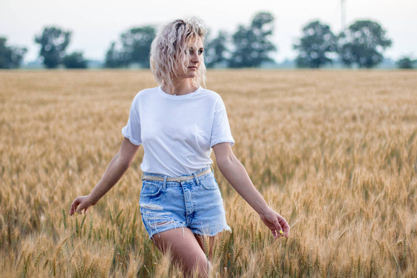 young tanned curly haired woman in white t-short and denim shorts on the field of wheat, wind turbine on the background - Photo, image