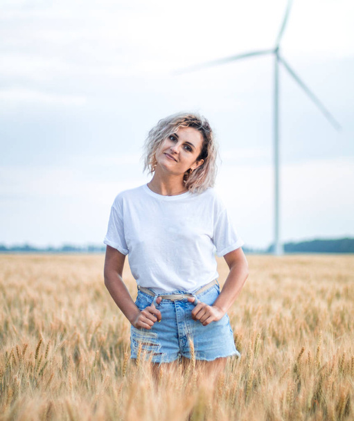 beyaz t-kısa ve kot şort genç tabaklanmış kıvırcık saçlı kadın olgunlaşmış buğday alanında olmaktan mutlu, arka planda rüzgar türbini - Fotoğraf, Görsel