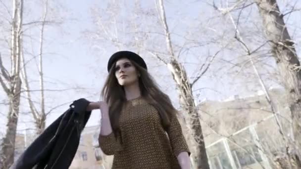 Νεαρό κορίτσι μοντέλο φορώντας καφέ φόρεμα και μαύρο καπέλο και το περπάτημα σε ένα πάρκο της πόλης με γυμνά δέντρα στο γαλάζιο του ουρανού φόντο, κάτω όψη. Δράση. Όμορφη γυναίκα που περπατάει έξω. - Πλάνα, βίντεο