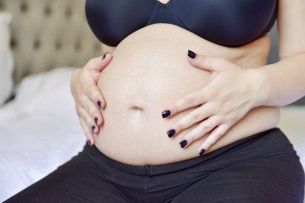 Primer plano de la mujer embarazada sentada en el sofá con las manos en el vientre
 - Foto, Imagen