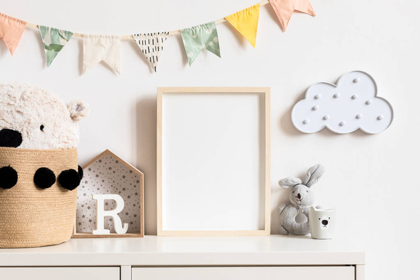 Stylish and modern scandinavian newborn baby interior with mock up photo or poster frame on the white shelf. Toys, teddy bear, plush rabbit and hanging cotton colorful flags and star. Template. Blank. - Photo, image