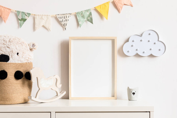 Stylish and modern scandinavian childroom interior with mock up photo or poster frame on the white shelf. Toys, teddy bear in basket, rocking horse and hanging cotton colorful flags and cloud. - Foto, afbeelding