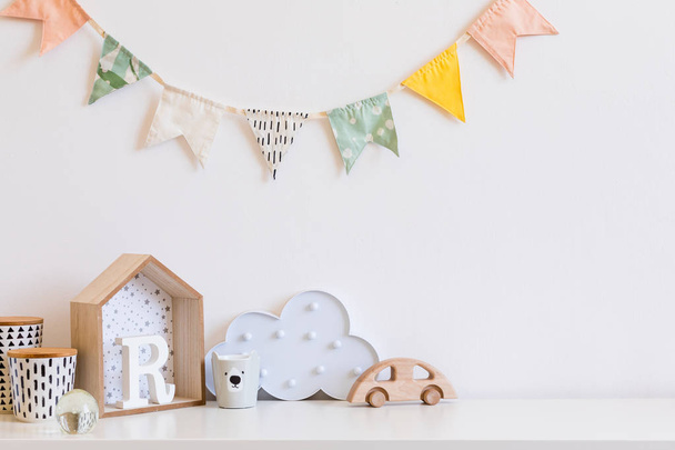 Stylish scandinavian nursery interior with wooden toys,boxes, accessories and cloud. Hanging colorful cotton flags on the white background wall. Cozy and sunny childroom. Copy space. - Foto, Imagen