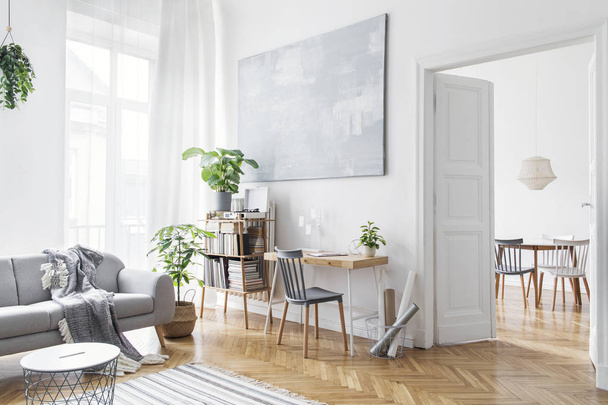 Stylish scandinavian open space with design furniture, plants, bamboo bookstand and wooden desk. Brown wooden parquet. Abstract painting. Modern decor of bright room next to dining room.  - Valokuva, kuva