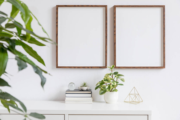 Stylish home interior with two brown wooden mock up photo frames on the white shelf with books, beautiful plants, gold pyramid and home accessories. Minimalistic concept of white room decor.  - Photo, image