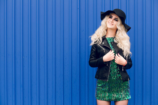 The girl in the black hat and leather jacket is standing and posing against the background of the blue wall - Foto, Imagem