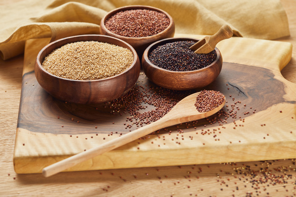 white, black and red quinoa in wooden bowls and spoon on wooden chopping board near napkin - Photo, Image