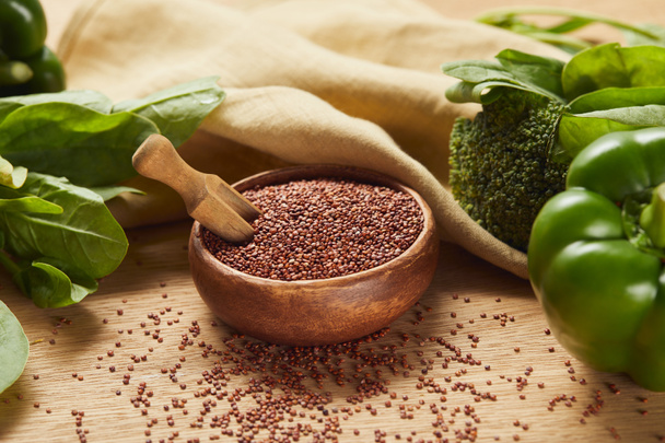 rode quinoa in houten kom met spatel in de buurt van beige servet en groene groenten - Foto, afbeelding