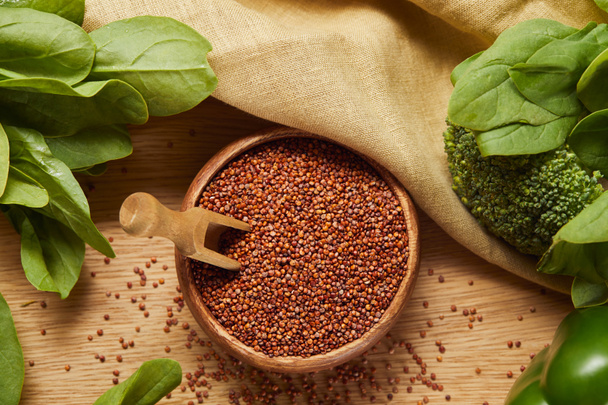 Ansicht von rotem Quinoa in Holzschale mit Spachtel in der Nähe von beiger Serviette und grünem Gemüse - Foto, Bild
