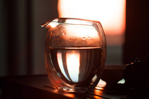 Glass pitcher of hot tea on wooden table on the background of beautiful sunset with sun rays. - Zdjęcie, obraz