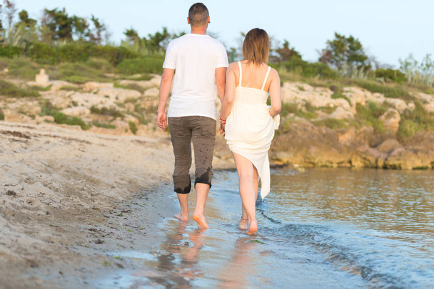 Υπαίθρια βολή από ρομαντικό νεαρό ζευγάρι που περπατά κατά μήκος της θάλασσας ακτή κρατώντας τα χέρια. Νέος άντρας και γυναίκα που περπατούν στην παραλία μαζί στο ηλιοβασίλεμα, κοντινό σώμα - Φωτογραφία, εικόνα