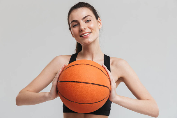 Young sport fitness woman player make exercises play basketball isolated over grey wall background. - Foto, afbeelding