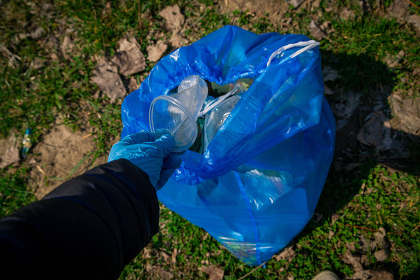 garbage in the nature, cleaning the environment in the spring on the river from the rubbish in disposable latex  mittens in to blue large plastic bag - Photo, Image
