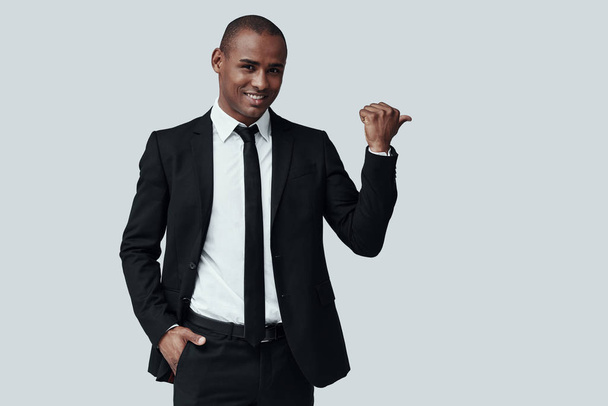 Take a look! Handsome young African man in formalwear looking at camera and pointing copy space while standing against grey background - Photo, Image