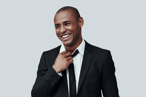 Werkdag is voorbij. Charmante jonge Afrikaanse man in formalwear aanpassen stropdas en glimlachen terwijl staande tegen grijze achtergrond - Foto, afbeelding
