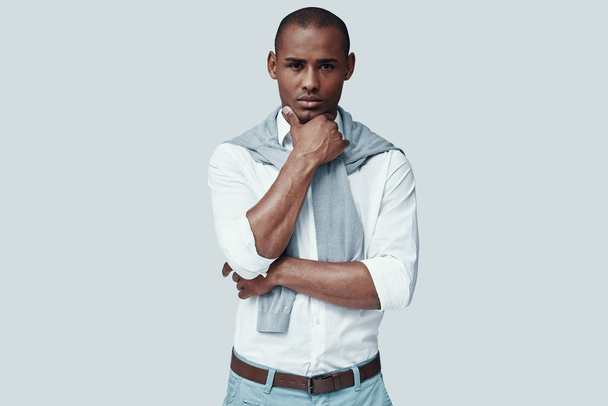 Thinking about... Handsome young African man looking at camera and keeping hand on chin while standing against grey background - Photo, Image
