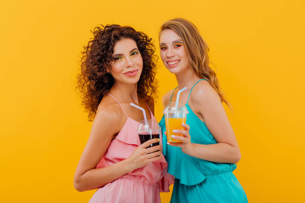 fashionable two cheerful girls with curly hair, hold the plastic cup with juice, look at the camera positive facial emotions, isolated on yellow background, copy space - Foto, imagen