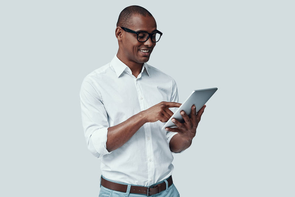  Examining new tablet. Handsome young African man using digital tablet and smiling while standing against grey background  - Photo, Image