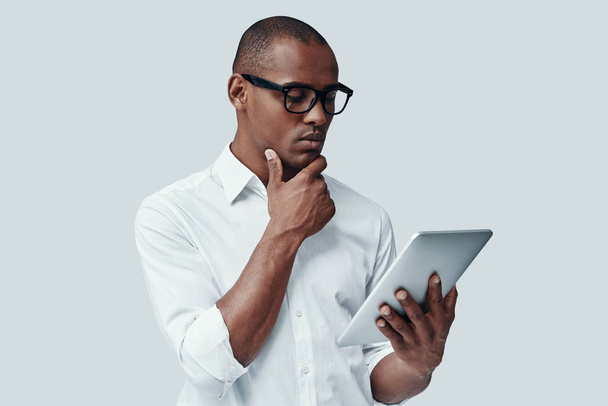 ¿Y si... Hombre africano joven guapo usando tableta digital y manteniendo la mano en la barbilla mientras está de pie contra el fondo gris
 - Foto, Imagen