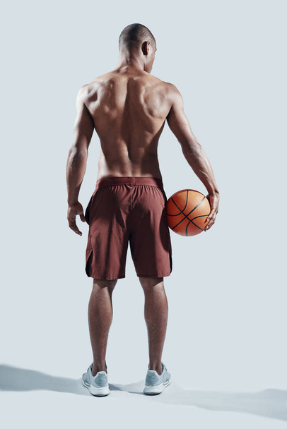 in großartiger Form. Rückansicht in voller Länge von hübschem jungen afrikanischen Mann in Sportkleidung, der Basketballball trägt, während er vor grauem Hintergrund steht - Foto, Bild