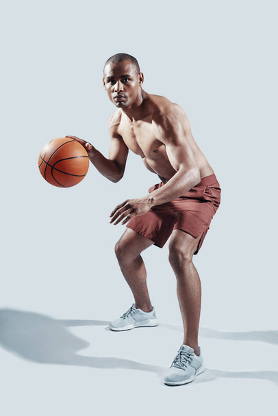 Inspanning te leveren. Volledige lengte van knappe jonge Afrikaanse man in sportkleding kijken naar camera tijdens het spelen van basketbal tegen grijze achtergrond - Foto, afbeelding