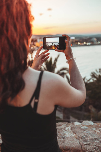 Beautiful woman taking a photo of sea landscape by mobile phone in the warm light of the sunset evening - Foto, Imagem