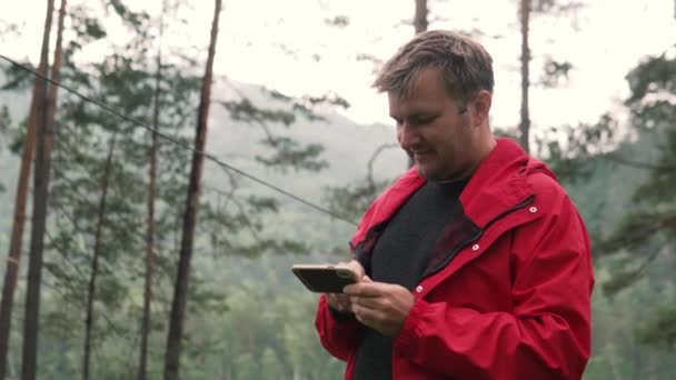 A man in a forest near a mountain river is looking at a mobile phone - Video