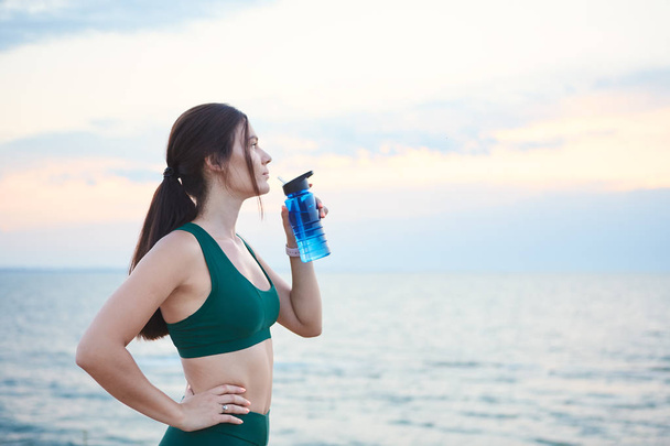 Young brunette woman with wireless earbuds and smart watches resting after morning workout drinking water. - Photo, image