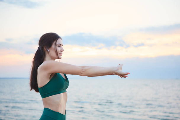 Mladá brunetka, která cvičává na mořském pobřeží při východu slunce a naslouchá hudbě. - Fotografie, Obrázek