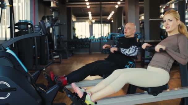 muscular man and fit woman do simultaneous seated cable rows on black machines in modern gym slow motion - Video, Çekim