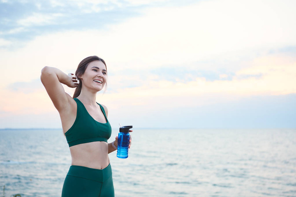 Young brunette woman with wireless earbuds and smart watches resting after morning workout drinking water. - Photo, Image
