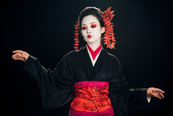 beautiful geisha in black and red kimono and flowers in hair looking down and gesturing isolated on black - Photo, Image