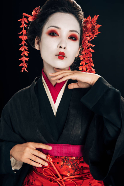 beautiful geisha in black and red kimono and flowers in hair touching chin isolated on black - 写真・画像