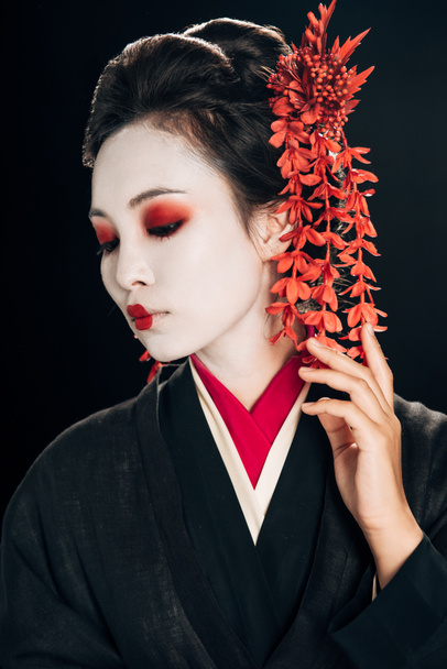 πορτρέτο της όμορφης γκέισα με μαύρο και κόκκινο κιμονό και λουλούδια στα μαλλιά κοιτάζοντας κάτω απομονωμένη σε μαύρο - Φωτογραφία, εικόνα