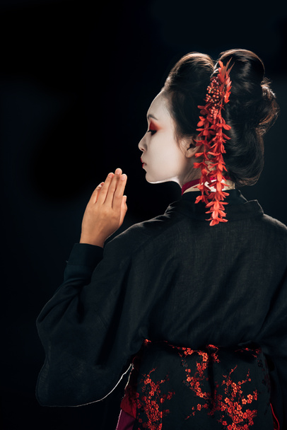 back view of beautiful geisha in black and red kimono and flowers in hair isolated on black - Foto, Imagem