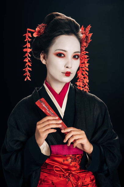 красивая гейша в черном кимоно с красными цветами в волосах держа традиционный ручной вентилятор изолирован на черном
 - Фото, изображение