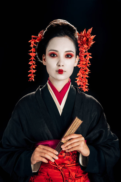 beautiful geisha in black and red kimono and flowers in hair holding wooden hand fan isolated on black - Photo, Image