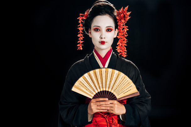 geisha in black kimono with red flowers in hair holding traditional asian hand fan isolated on black - Фото, изображение