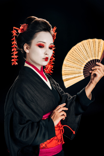 gueixa em quimono preto com flores vermelhas no cabelo segurando tradicional asina ventilador de mão isolado em preto
 - Foto, Imagem