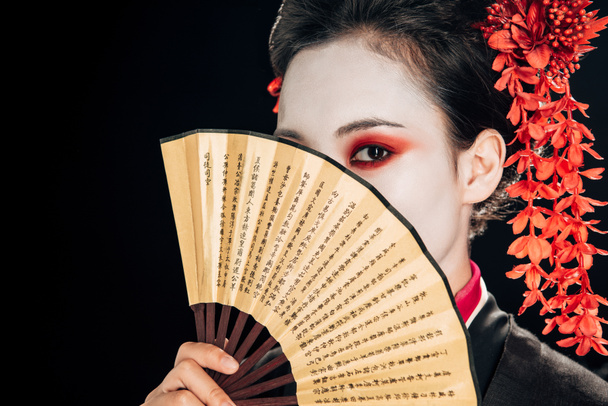geisha joven en kimono negro con flores rojas en el pelo sosteniendo ventilador de mano tradicional en frente de la cara aislado en negro
 - Foto, Imagen