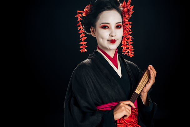happy geisha in black kimono with red flowers in hair holding traditional asian hand fan isolated on black - Zdjęcie, obraz