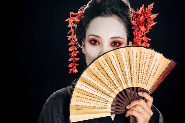 geisha in black kimono with red flowers in hair holding traditional asian hand fan near face isolated on black - Фото, изображение