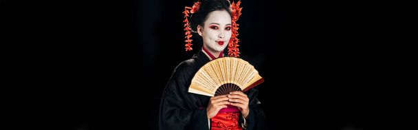 χαμογελαστή γκέισα με μαύρο κιμονό με κόκκινα λουλούδια στα μαλλιά κρατώντας παραδοσιακό ανεμιστήρα χεριού απομονωμένο σε μαύρο, πανοραμικό σουτ - Φωτογραφία, εικόνα