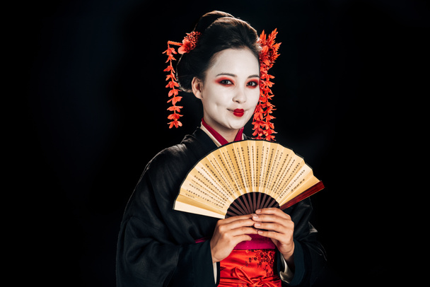 χαρούμενο γκέισα σε μαύρο κιμονό με κόκκινα λουλούδια στα μαλλιά που κατέχουν παραδοσιακό ασιατικό χέρι ανεμιστήρα απομονωθεί σε μαύρο - Φωτογραφία, εικόνα