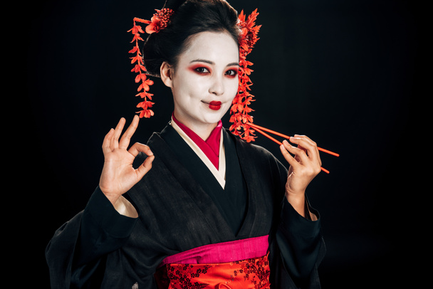 mosolygó szép gésa fekete kimonó vörös virágokkal a haj gazdaság pálcika és mutatja Oké jel elszigetelt fekete - Fotó, kép