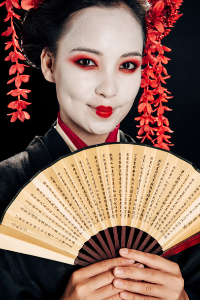 portrait of smiling beautiful geisha in black and red kimono and flowers in hair holding hand fan isolated on black - Photo, Image