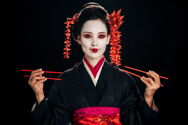 happy beautiful geisha in black kimono with red flowers in hair holding chopsticks isolated on black - Zdjęcie, obraz