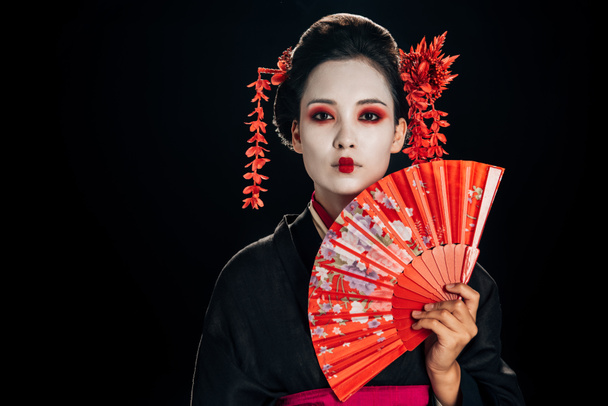 attractive geisha in black kimono with flowers in hair holding bright hand fan isolated on black with copy space - Photo, Image
