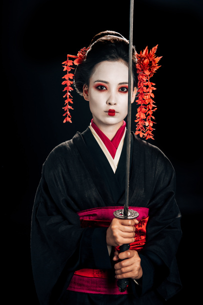 όμορφη γκέισα με μαύρο κιμονό με κόκκινα λουλούδια στα μαλλιά κρατώντας παραδοσιακή Κατάνα απομονωμένη σε μαύρο - Φωτογραφία, εικόνα