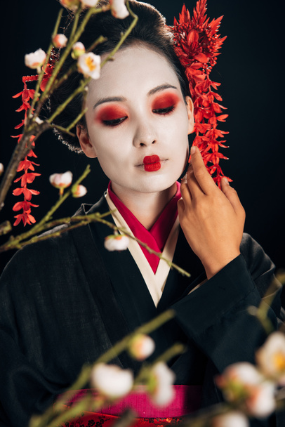 selective focus of beautiful geisha in black kimono with red flowers in hair touching face and sakura branches isolated on black - Zdjęcie, obraz